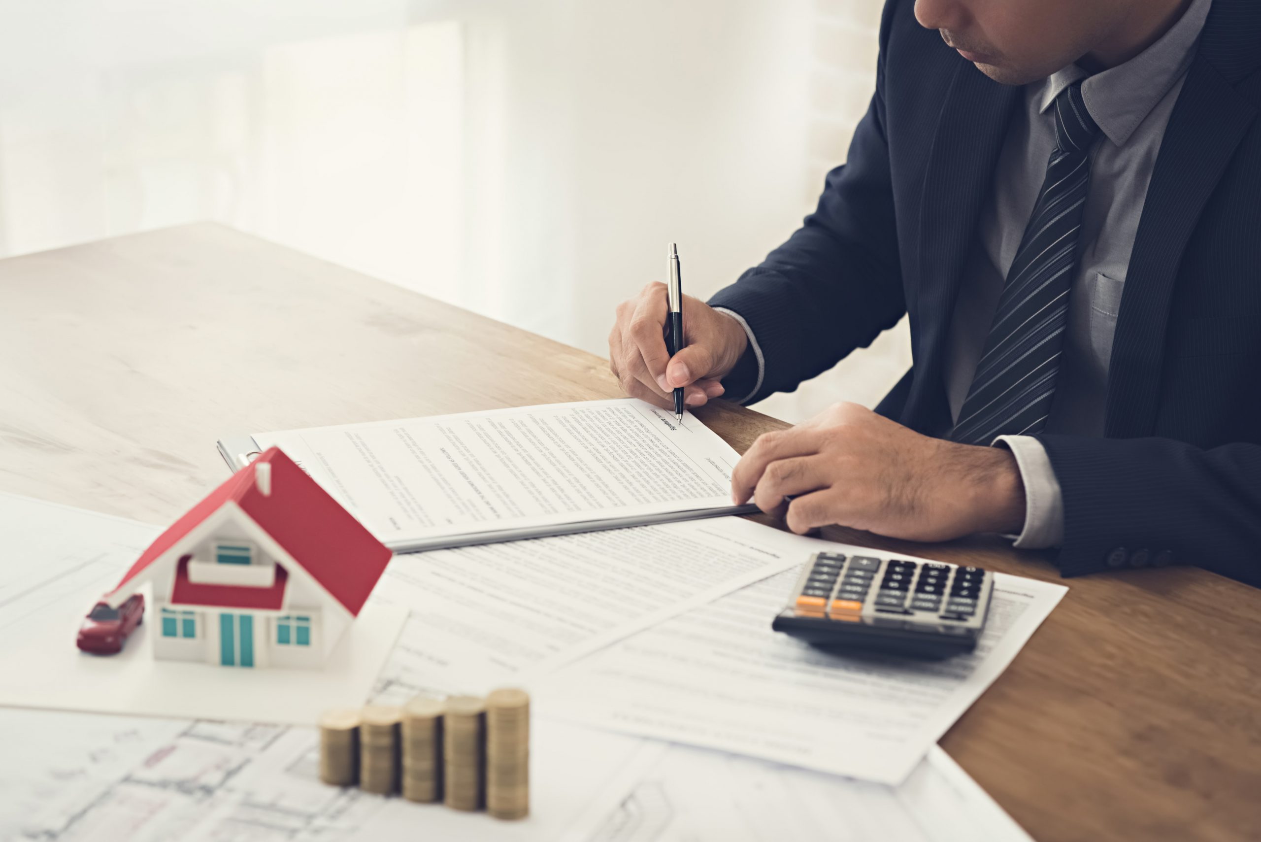 Pictured is a man at the table with a pen in his hand, notebook on the table, calculator, coins, and a small house.